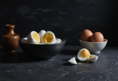 Halved boiled eggs on thick Greek yogurt in a dark bowl beside whole brown eggs, on dark slate under raking sidelight