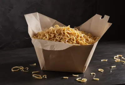 An open paper takeaway carton overfilled with noodles and vegetables heaped above its rim, sitting on a pale wooden counter with the lid propped beside it.