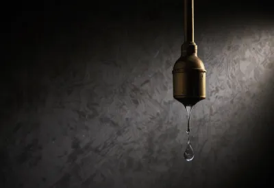 A single bright water drop falling from a weathered brass spout, lit by one hard spotlight against a dark background