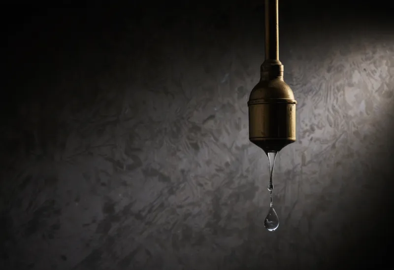 A single bright water drop falling from a weathered brass spout, lit by one hard spotlight against a dark background