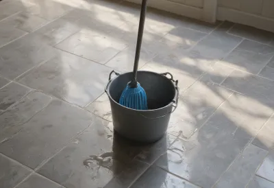 A metal mop bucket filled with grey water standing on a wet tiled kitchen floor, lit by low afternoon sun.