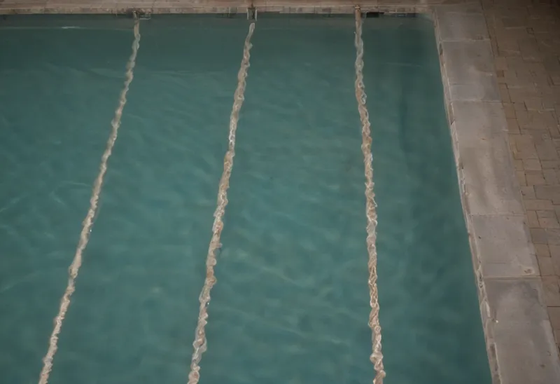 Overhead view of an empty floodlit swimming pool at night, its still surface divided into parallel stripes by lane ropes.