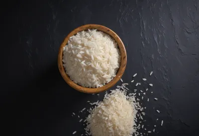 A plain wooden cup piled well above its rim with cooked white rice, grains spilling onto a dark slate surface