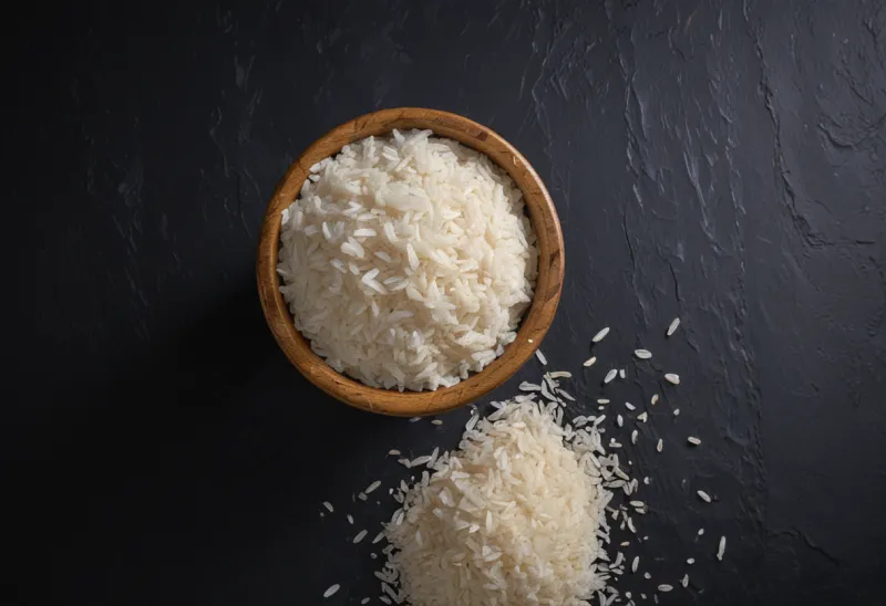 A plain wooden cup piled well above its rim with cooked white rice, grains spilling onto a dark slate surface