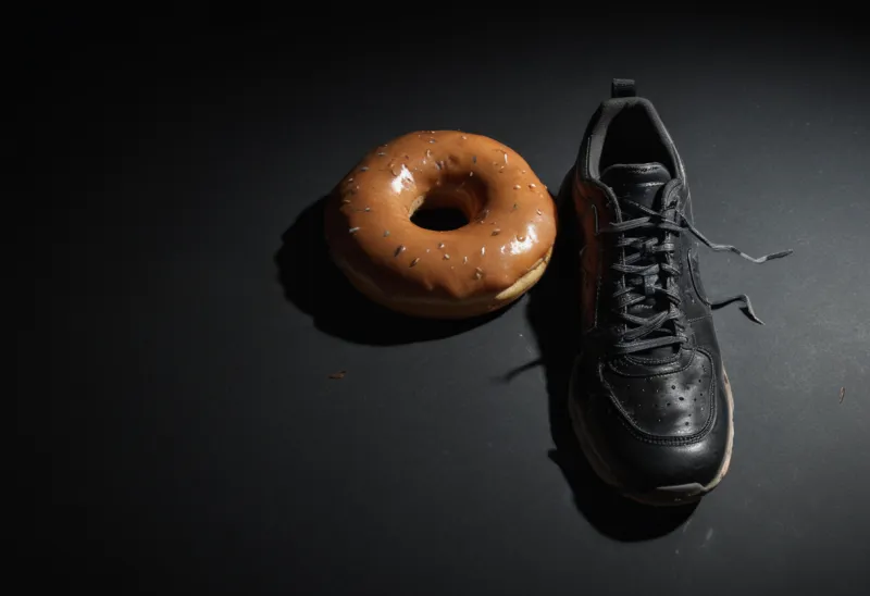 A single glazed doughnut next to one worn running shoe on a bare dark surface
