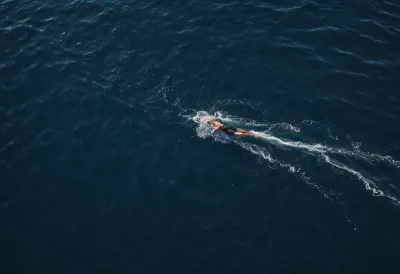 A lone swimmer seen from high above cutting across dark blue open water, a single clean wake trailing behind, small in the frame