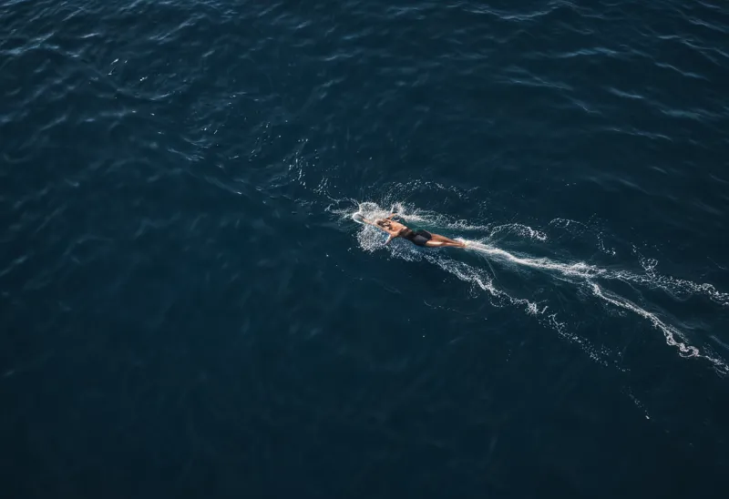 A lone swimmer seen from high above cutting across dark blue open water, a single clean wake trailing behind, small in the frame
