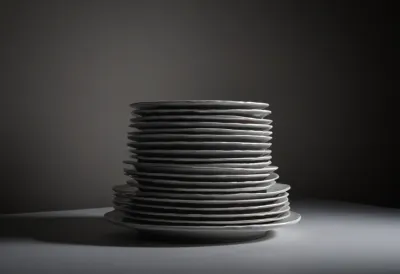 A tall stack of white dinner plates leaning noticeably off-vertical on a plain dark table