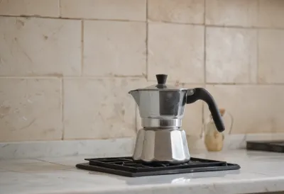 A battered aluminium moka pot venting steam on a pale tiled kitchen counter.