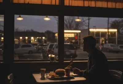 Silhouette of a person seated at a restaurant window at dusk facing a large plate of burger and fries, lit from behind