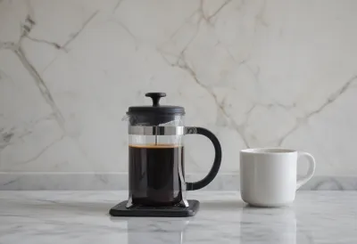 A single French press of black coffee on a marble counter with one empty white mug set to its left, in soft morning light