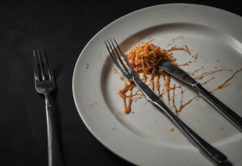 A fork and knife laid together diagonally across a used plate with faint sauce smears and a few crumbs, the meal just finished, against a dark background