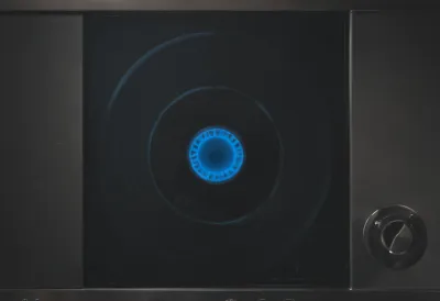 A gas hob burner turned to its lowest setting, showing a single thin unbroken ring of blue burning gas on a dark stovetop, photographed close from above.