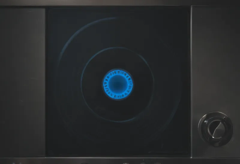 A gas hob burner turned to its lowest setting, showing a single thin unbroken ring of blue burning gas on a dark stovetop, photographed close from above.