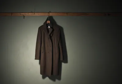 A single plain wool coat on a wooden hanger, hanging alone from a hook on a bare wall