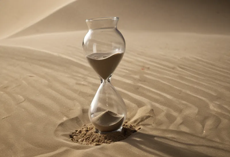 Close side view of a small hourglass in mid-flow, its upper bulb half drained and a thin stream of pale sand falling into the lower bulb.