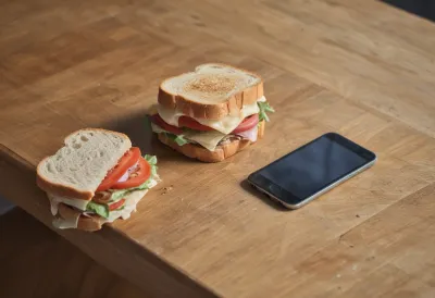 A phone face down on a wooden table next to a half-eaten sandwich on a plate