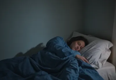 A sleeping figure under a rumpled duvet across a dim bedroom at blue hour, lit only by faint window light