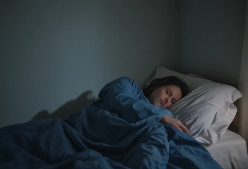A sleeping figure under a rumpled duvet across a dim bedroom at blue hour, lit only by faint window light