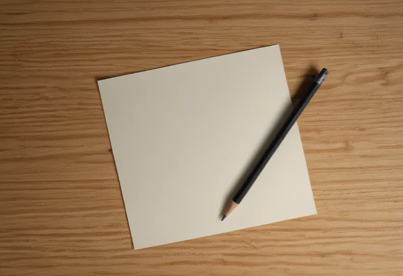 A graphite pencil lying across a blank cream note card on a plain oak table, seen from above.