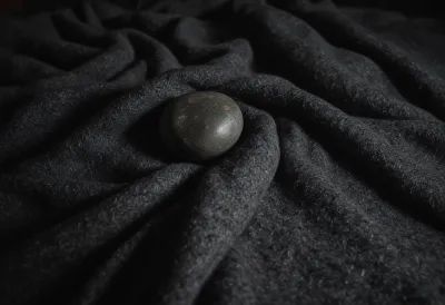 A dark river stone half sunk into a thick grey wool blanket under hard sidelight, the fabric dimpled deeply around it