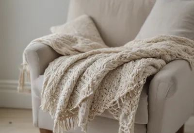 A folded knitted wool blanket draped over the arm of a linen armchair