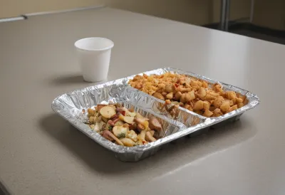 A half-eaten meal in a foil tray on a bare table in a windowless staff break room under flat fluorescent light.
