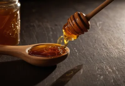 A slow, thick thread of amber honey trailing from a wooden dipper, caught mid-drip in warm morning light against deep shadow