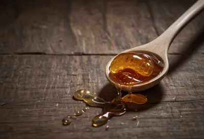A heaped spoon of honey on pale weathered wood with a slow amber thread running off its edge