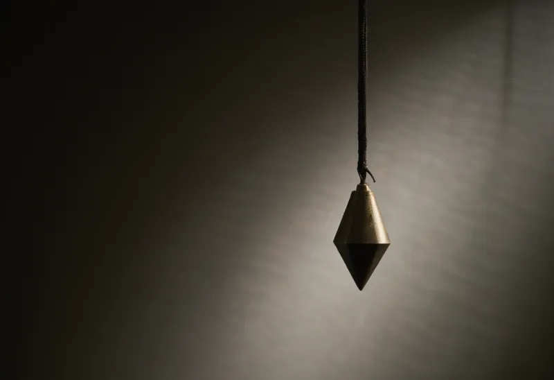 A single brass plumb bob hanging motionless and centered on a thin cord, lit by one hard shaft of light against deep shadow